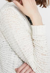 Osoba v bílém pleteném svetru s texturovanými otvorenými stehy, ruka položená blízko brady, částečně viditelný květinový oděv pod ním.