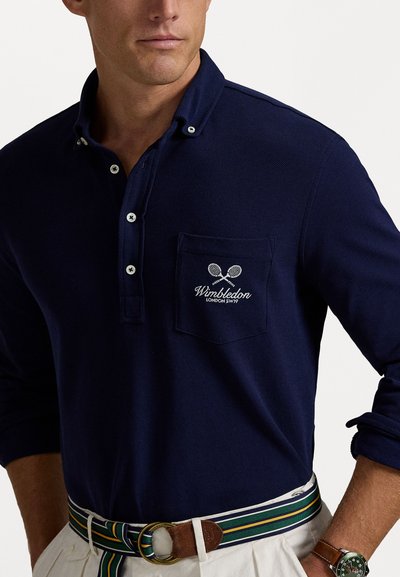 Navy long-sleeve polo with a buttoned collar, featuring an embroidered Wimbledon logo on the chest pocket and ribbed texture. Styled with a striped belt.