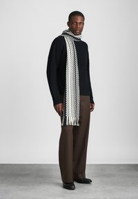 Pletený čierno-biely šál so vzorom zigzag, s třásněmi na koncoch. Nosený cez tmavý sveter a hnedé nohavice.