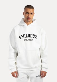 Sweat à capuche blanc avec une poche avant et des poignets côtelés, arborant "SMILODOX ATHL. WEAR" en lettres noires sur la poitrine.