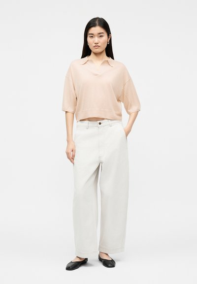 Femme aux cheveux noirs et lisses portant un haut beige ample à manches courtes, un pantalon large blanc et des chaussures plates noires, debout devant un fond uni.