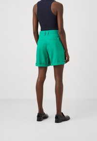 Grüne Shorts mit glatter Textur, umgeschlagenem Saum und Gesäßtaschen, kombiniert mit einem figurbetonten schwarzen ärmellosen Oberteil und schwarzen Schuhen.