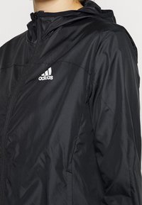 Persona che indossa una giacca con cappuccio nera di Adidas, con una cerniera frontale e il logo bianco di Adidas sul petto, mostrata dalla spalla alla vita.