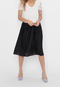 Gonna midi plissettata nera con una texture liscia. Abbinata a un top bianco in pizzo a maniche corte e tacchi lavanda, che mette in risalto un look elegante.