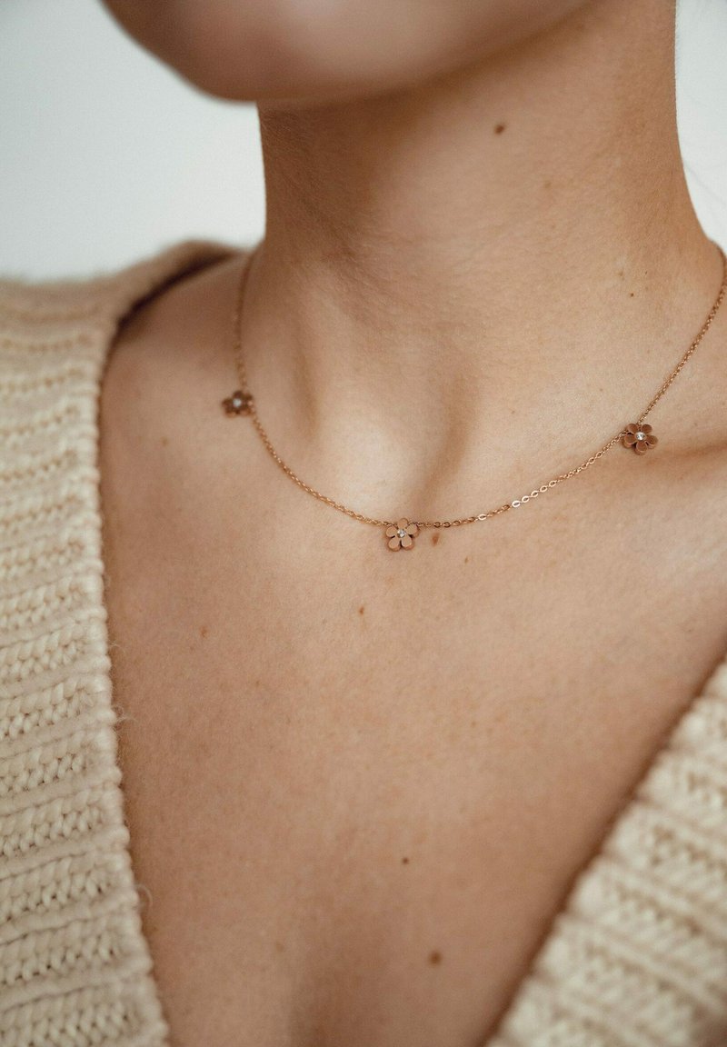 Collana a forma di fiore in oro rosa con piccoli accentuati floreali su una catena delicata, su uno sfondo di maglione leggero e texturizzato.