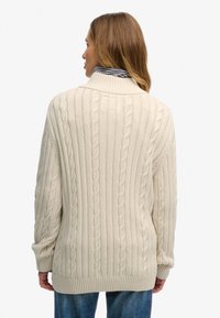 Pull beige en maille torsadée avec un col montant, des poignets et un ourlet côtelés. La texture présente des motifs en relief, offrant une apparence chaleureuse.