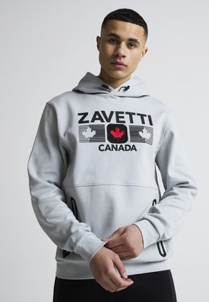 Jeune homme portant un sweat à capuche gris clair avec le texte "ZAVETTI CANADA" et un logo d'érable rouge sur le devant, debout devant un fond uni.