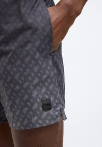 Sive kopalne hlače z vzorcem iz mešanice geometrijskih oblik. Imajo stranski žep in črno pravokotno etiketo z logotipom na robu.