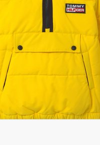 Żółta kurtka puchowa z zamkiem z przodu, przednią kieszenią z klapką i zatrzaskiem oraz czarną naszywką z logo. Gładki, izolowany materiał.