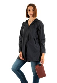 Chubasquero azul marino resistente al agua con capucha, cierre a presión, dos bolsillos laterales y un corte holgado, combinado con un neceser cosmético marrón.