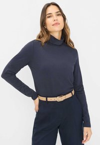 Navy langærmet turtleneck skjorte lavet af blødt stof, med et tætsiddende design, kombineret med højderetstående mørke bukser og et beige bælte.
