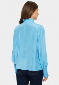 Blusa azzurra chiara in tessuto lucido, con colletto alto arricciato, maniche raccolte e una vestibilità comoda con orlo leggermente curvo.