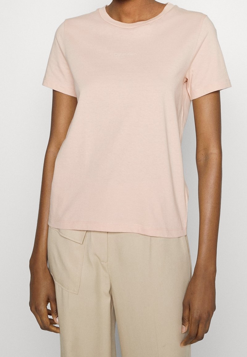Light pink cotton t-shirt with a round neckline and short sleeves. Features a subtle logo in the center. Worn with beige pants.