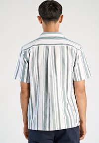 Man in een wit shirt met korte mouwen en verticale grijze strepen, met de rug naar de kijker, tegen een effen witte achtergrond.