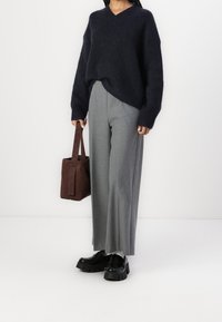 Personne portant un pull oversize foncé, un pantalon gris à jambe large, des chaussures noires épaisses et tenant un sac fourre-tout en daim marron sur fond blanc.