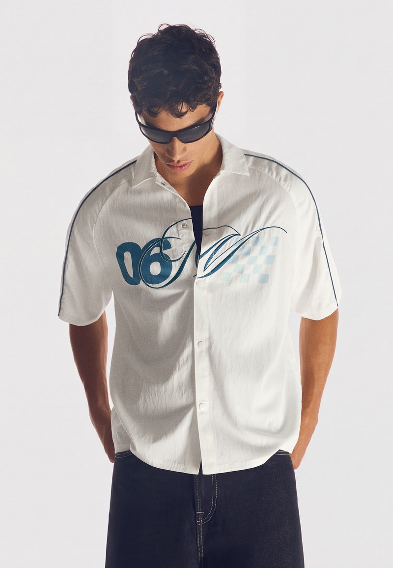Homme portant des lunettes de soleil noires, chemise blanche à manches courtes avec un motif bleu et pantalon foncé, regardant vers le bas, les mains dans le dos.