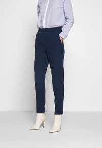 Person i marinblå snäva byxor, blå och vit randig skjorta med knappar, och vita högklackade ankelskor, står mot en enfärgad bakgrund.