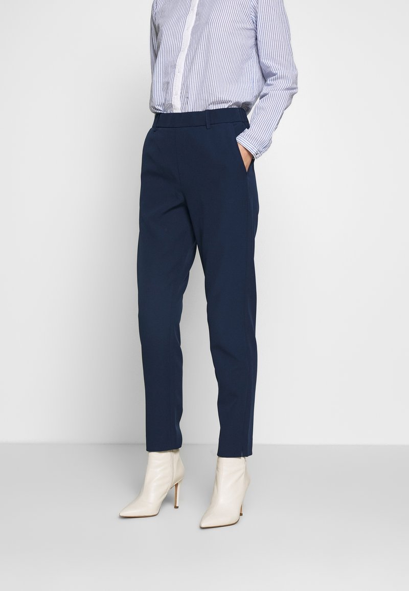 Person i marinblå snäva byxor, blå och vit randig skjorta med knappar, och vita högklackade ankelskor, står mot en enfärgad bakgrund.