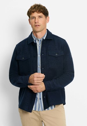 Marineblauer Wollmischungsjacke mit zwei Brusttaschen, Knopfverschluss und strukturierter Oberfläche, über einem blau-weißen gestreiften Hemd getragen.