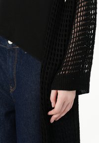 Cardigan preto em tricot aberto com um padrão solto, combinado com uma blusa preta justa e jeans escuros de lavagem azul. Colocação de mão notável na manga.