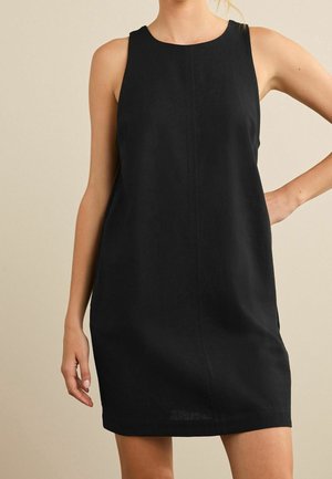 Woman wearing sleeveless, black knee-length dress with a round neckline against a plain beige background.