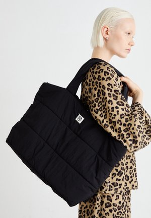 Woman with short blonde hair wearing leopard print outfit carries large black quilted tote bag over shoulder.