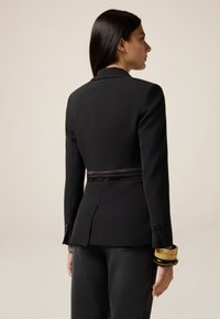 Vrouw in een aansluitende zwarte blazer met geplooide details aan de achterkant en brede zwarte en gouden armbanden, staand tegen een effen achtergrond.