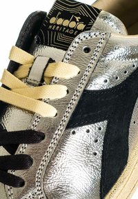 Close-up van een metallic zilveren en zwarte sneaker met crèmekleurige en zwarte veters, geperforeerde details en het "diadora HERITAGE" logo op de tong.