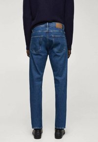 Vaqueros de mezclilla azul oscuro con corte recto, con bolsillos traseros y un parche de cuero marrón en la cinturilla. Llevados con zapatos negros.