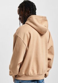 Smėlinė per didelė džemperis su raišteliu ant kapučino, sutrauktomis rankovėmis ir apačioje. Minkšta tekstūra, paprastas dizainas, matoma iš nugaros, derinama su mėlynomis džinsomis.