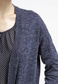 Mörkblå stickad cardigan med öppen front, texturerad tyg, långa ärmar och avslappnad passform, lager över en blus med prickar.