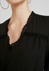 Blouse noire avec un col en V orné d'une dentelle raffinée. Tissu doux avec un léger éclat, détails plissés sous les accents en dentelle.