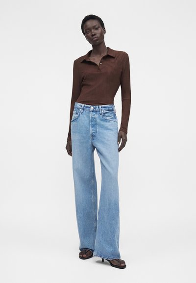 Γυναίκα που φοράει στενή καφέ ριμπ πουλόβερ πόλο με μακριά μανίκια, ανοιχτό μπλε φαρδιά τζιν με ψηλή μέση και μαύρα ανοιχτά πέδιλα.