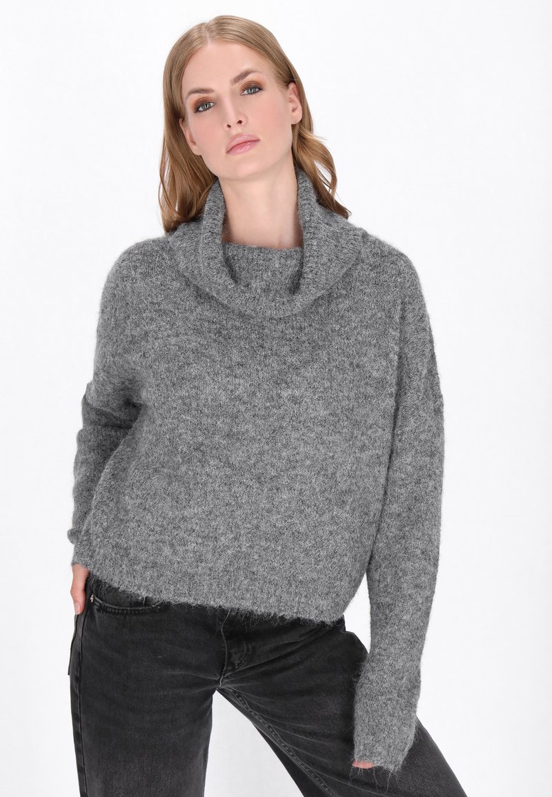 Frau in einem grauen, flauschigen Rollkragenpullover und schwarzen Jeans, die vor einem weißen Hintergrund posiert.