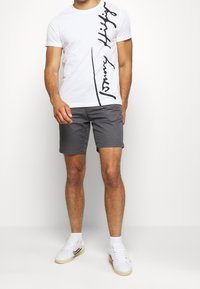 Camiseta de algodón blanca con el logo de "Tommy Hilfiger" escrito a mano en negro. Pantalones cortos gris oscuro de corte recto. Zapatillas blancas con acentos rojos y azules.