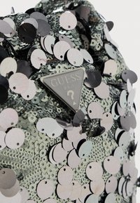 Paillettes argentate lucenti coprono densamente il tessuto, con una placca in metallo triangolare del logo Guess U.S.A. centrata tra le paillettes.