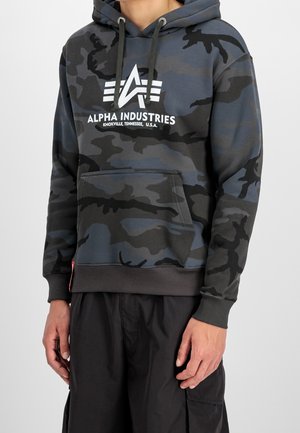 Homme portant un sweat à capuche camouflage noir et gris avec le logo « Alpha Industries Knoxville Tennessee U.S.A. » et un pantalon cargo noir.