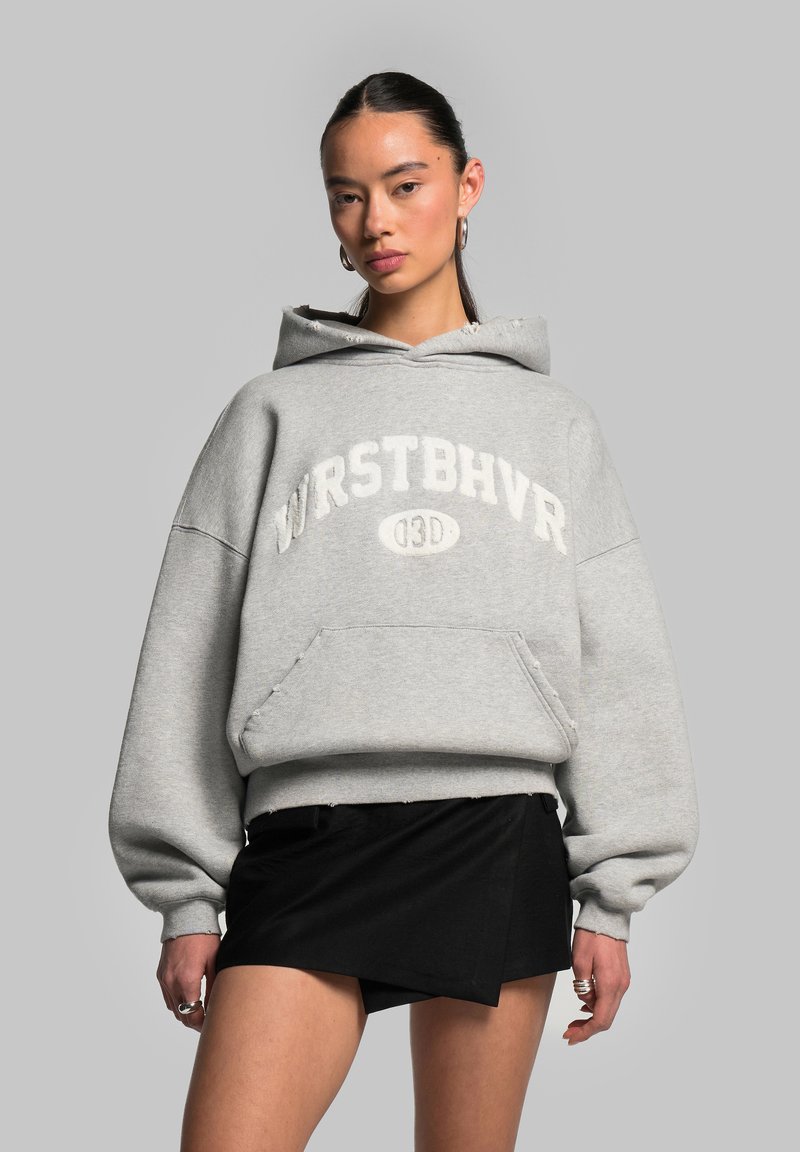 Felpa grigia oversize con il testo "WRSTBHVR" e tasca a marsupio. Presenta un orlo grezzo e un tessuto texturizzato. Abbinata a una mini gonna nera.
