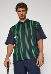Korte ermer, marineblå mesh polo skjorte med grønne vertikale striper, krage og halv glidelås. Har logodetalj på venstre side.