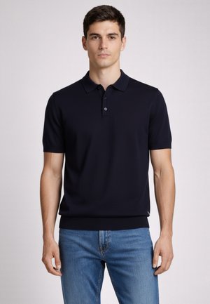 Młody mężczyzna w jednolitej czarnej, krótkim rękawem koszulce polo i niebieskich dżinsach, stojący na tle jednolitego jasnego tła.