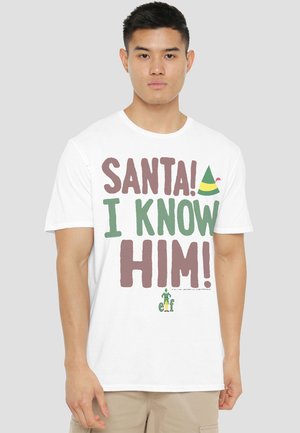 Young man wearing a white t-shirt with "SANTA! I KNOW HIM!" text and a small elf hat graphic on a plain light gray background.