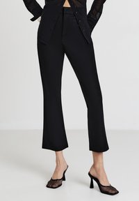 Donna che indossa pantaloni neri a vita alta, una camicia nera trasparente con bottoni e mule nere in rete con tacco alto, su uno sfondo bianco.