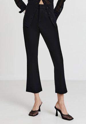 Donna che indossa pantaloni neri a vita alta, una camicia nera trasparente con bottoni e mule nere in rete con tacco alto, su uno sfondo bianco.