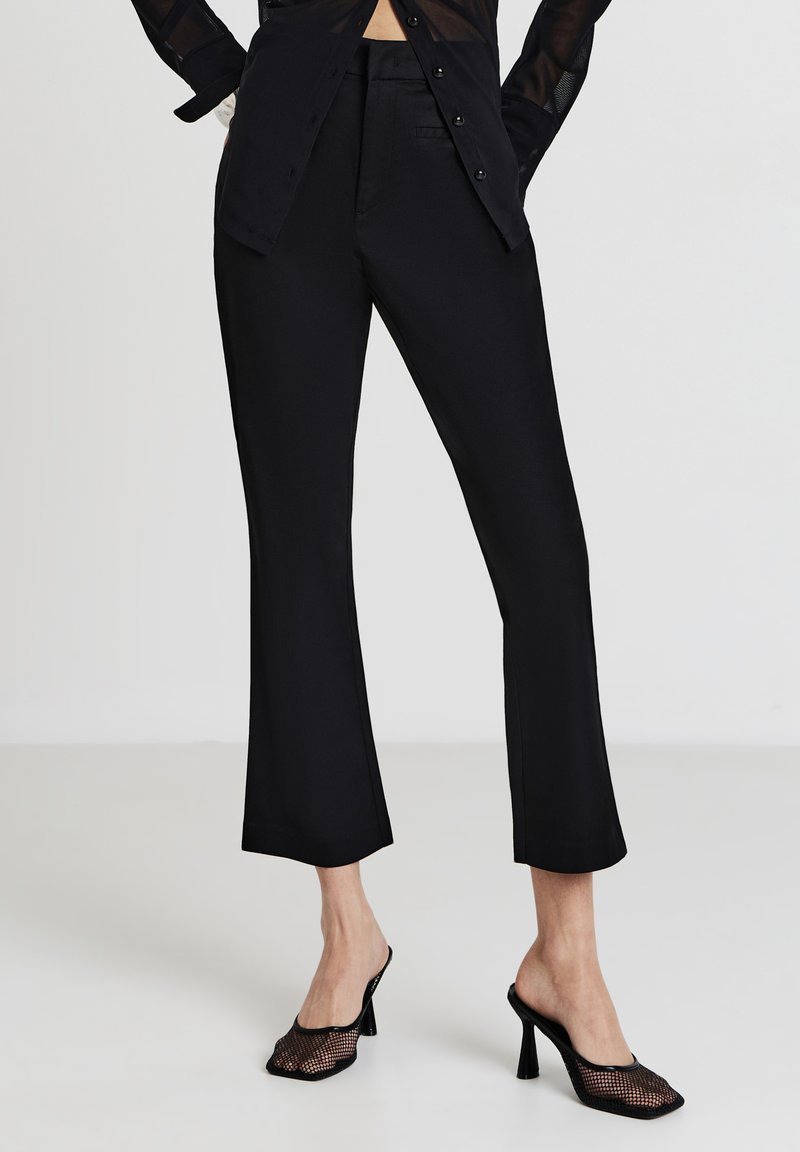 Donna che indossa pantaloni neri a vita alta, una camicia nera trasparente con bottoni e mule nere in rete con tacco alto, su uno sfondo bianco.