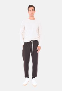 Uomo in piedi con indosso un maglione bianco a maniche lunghe, pantaloni neri con cordino, calzini neri e sneakers bianche su uno sfondo neutro.