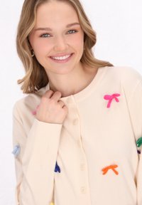 Cardigan bej deschis cu un închizător cu buton, având fundițe din țesătură colorată în diverse nuanțe cusute pe față și pe mâneci.