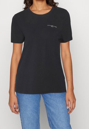 T-shirt en coton noir à manches courtes, col rond, avec un logo blanc imprimé sur le côté gauche de la poitrine, associé à un jean en denim bleu clair.