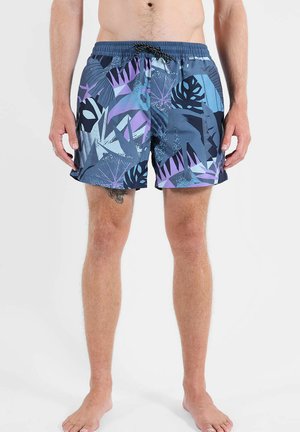 Homme portant un short de bain à imprimé tropical bleu et violet avec une taille élastique et un cordon, debout pieds nus sur un fond blanc.