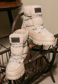 Stivali beige foderati in pelliccia con suola robusta, caratterizzati da una tomaia testurizzata, lacci funzionali e un'etichetta con logo sul lato esterno.