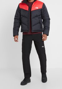Giacca a vento nera e rossa con design trapuntato, cerniere e logo. Abbinata a pantaloni neri con accenti e tasche. Stivali neri.